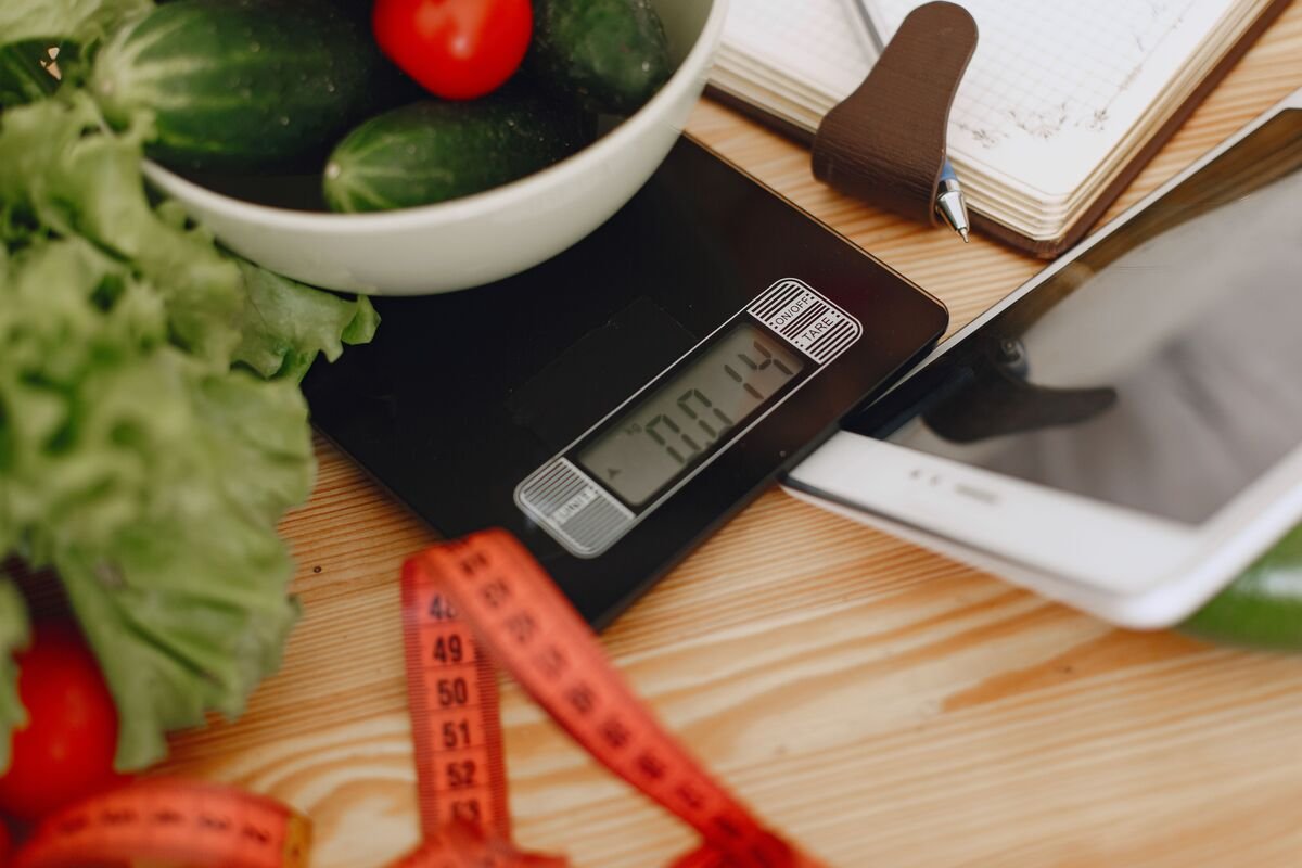 Digital kitchen scale with fresh vegetables, measuring tape and notebook for calorie tracking and diet planning